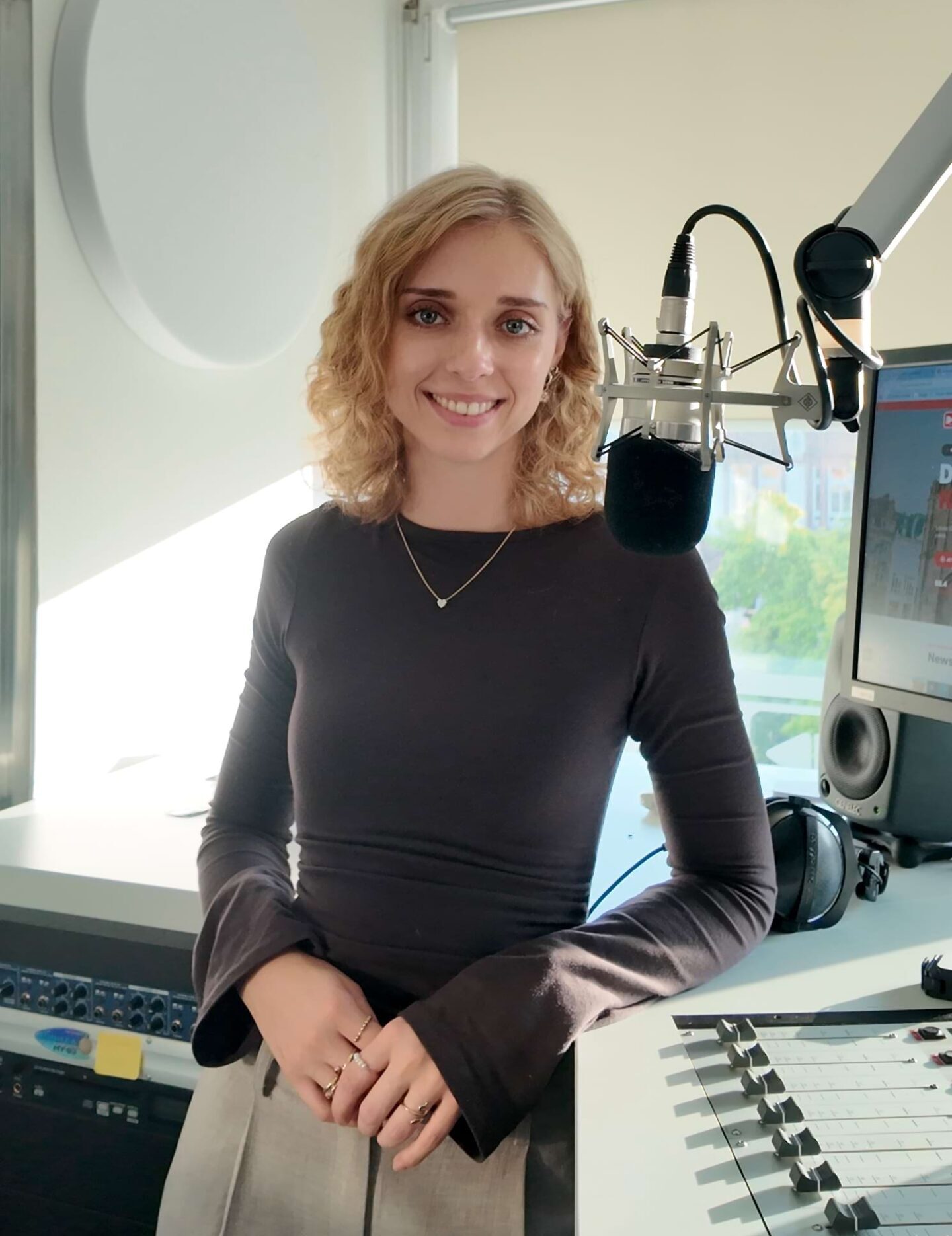Eine junge Frau mit lockigem, blondem Haar steht lächelnd vor einem Mikrofon in einem Radio-Studio.