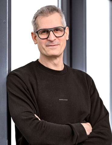 Ein Mann mit Brille und kurzem Haar, gekleidet in einem schwarzen Sweatshirt, lehnt entspannt an einer Wand.