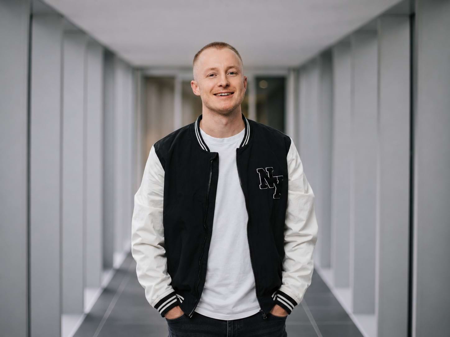Mann mit kurzem Haar und lächelndem Gesicht trägt eine schwarze und weiße Jacke und steht in einem modernen Gebäude.